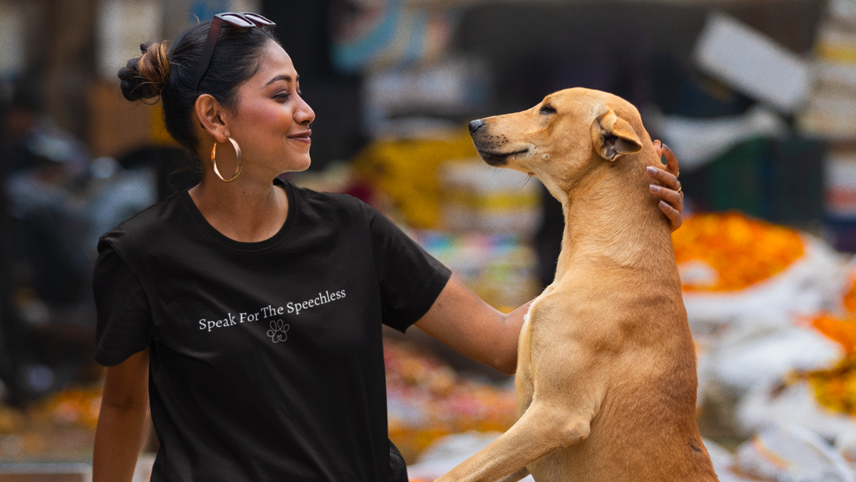 Woman sitting with a dog at a market stall selling flowers and fruits with a tshirt saying 'speak for the speechless'.