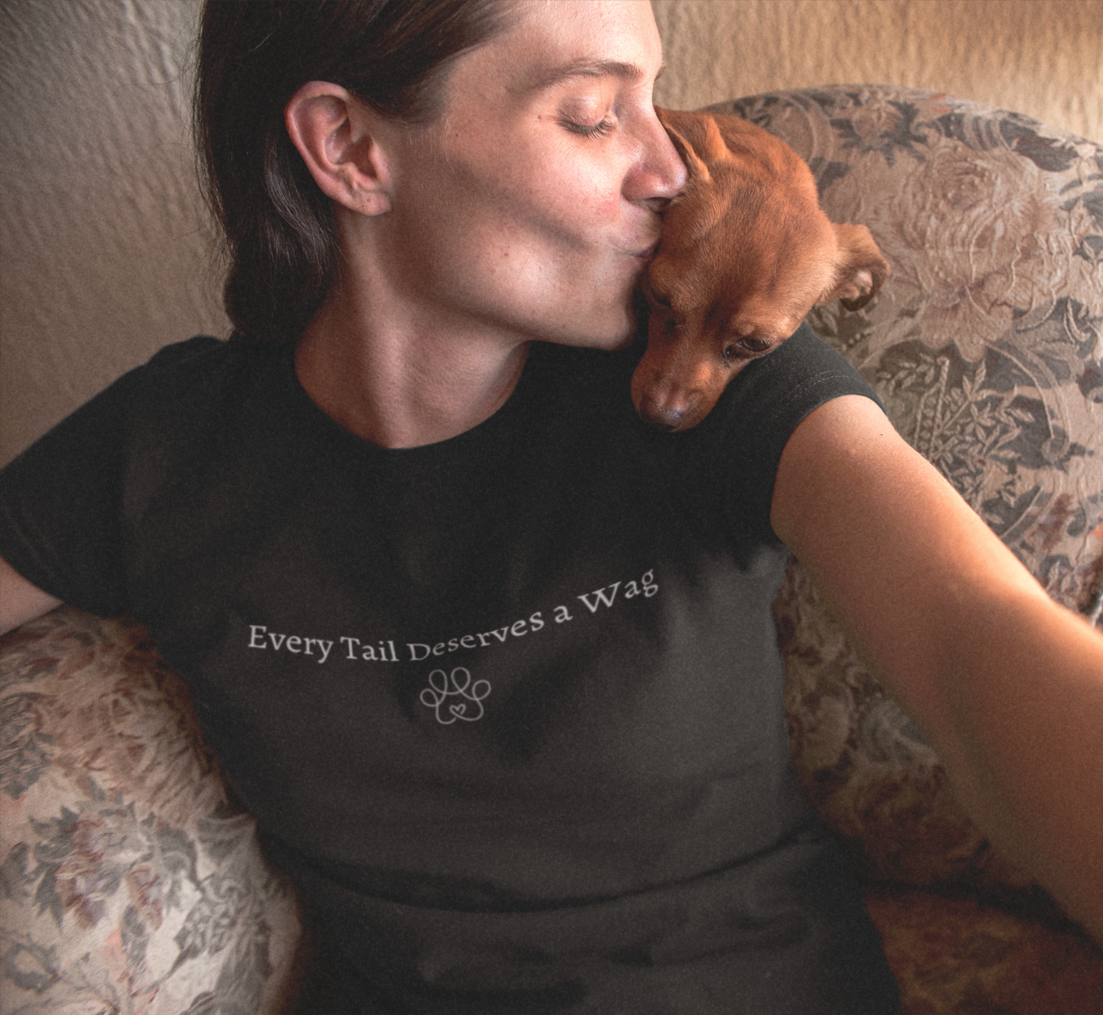 Person wearing a shirt with 'Every Tail Deserves a Wag' on a floral couch with a dog.