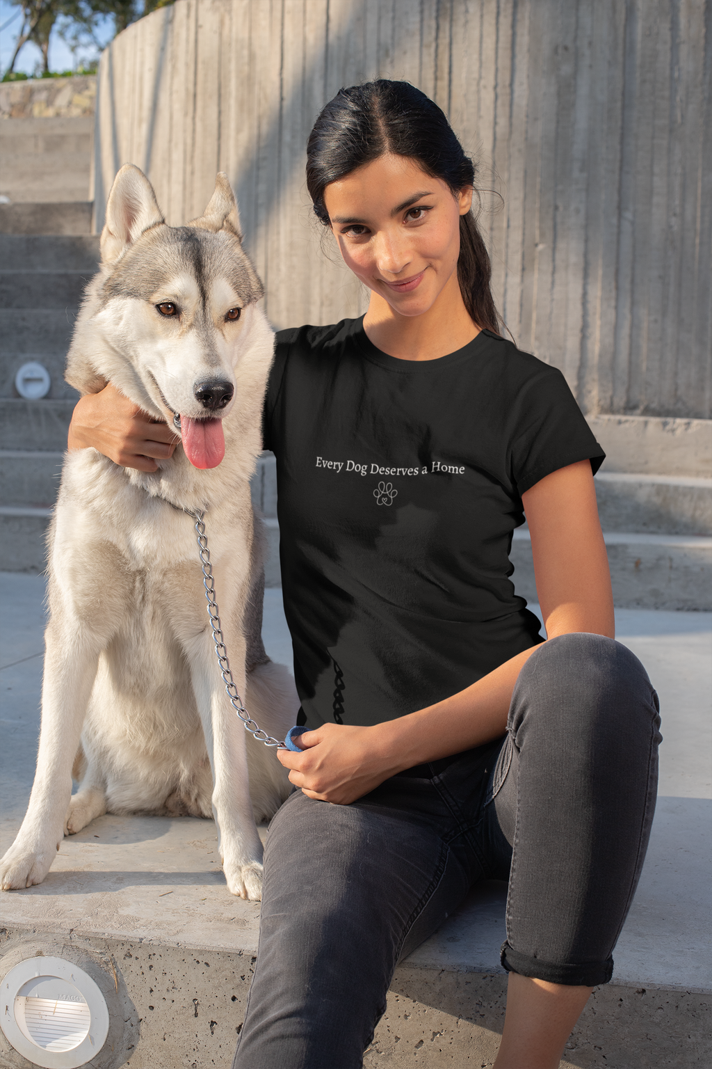 Woman sitting with a dog wearing a black t-shirt with 'every dog deserves a home' text, outdoors.